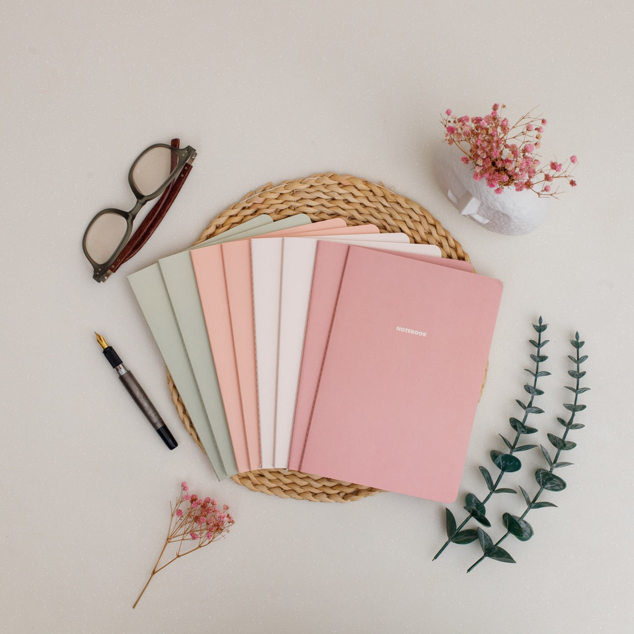 Set of colorful notebooks in a woven basket with a pen, glasses, and flowers on a light surface.