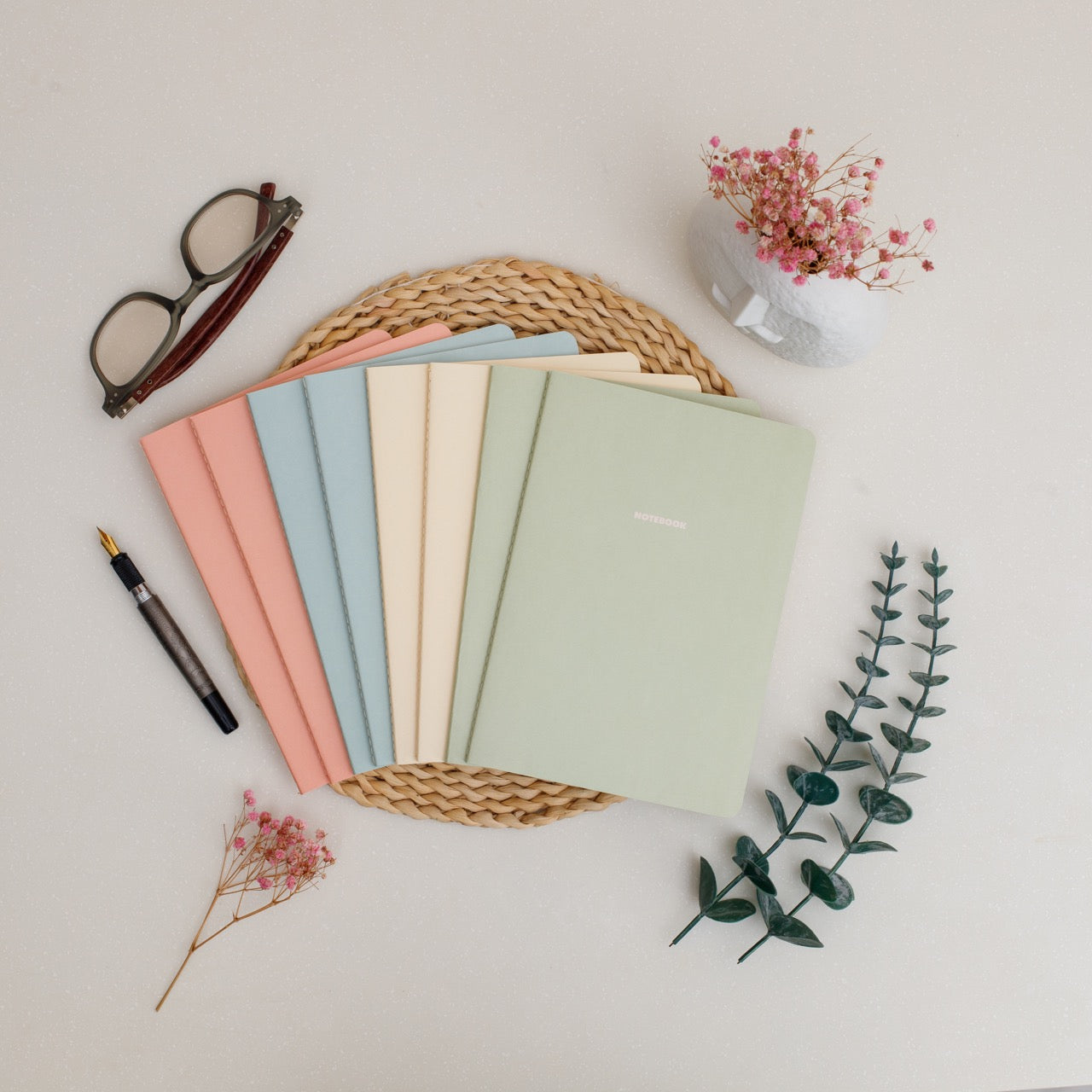 8 spring colored notebooks on a woven mat with a pen, glasses, and flowers on a light background