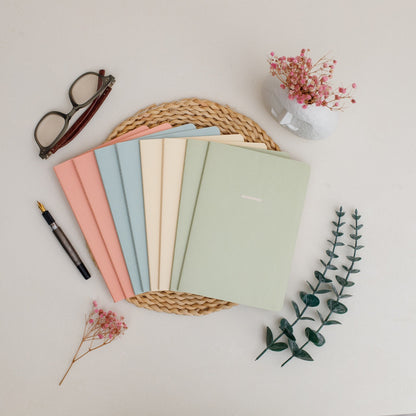 8 spring colored notebooks on a woven mat with a pen, glasses, and flowers on a light background