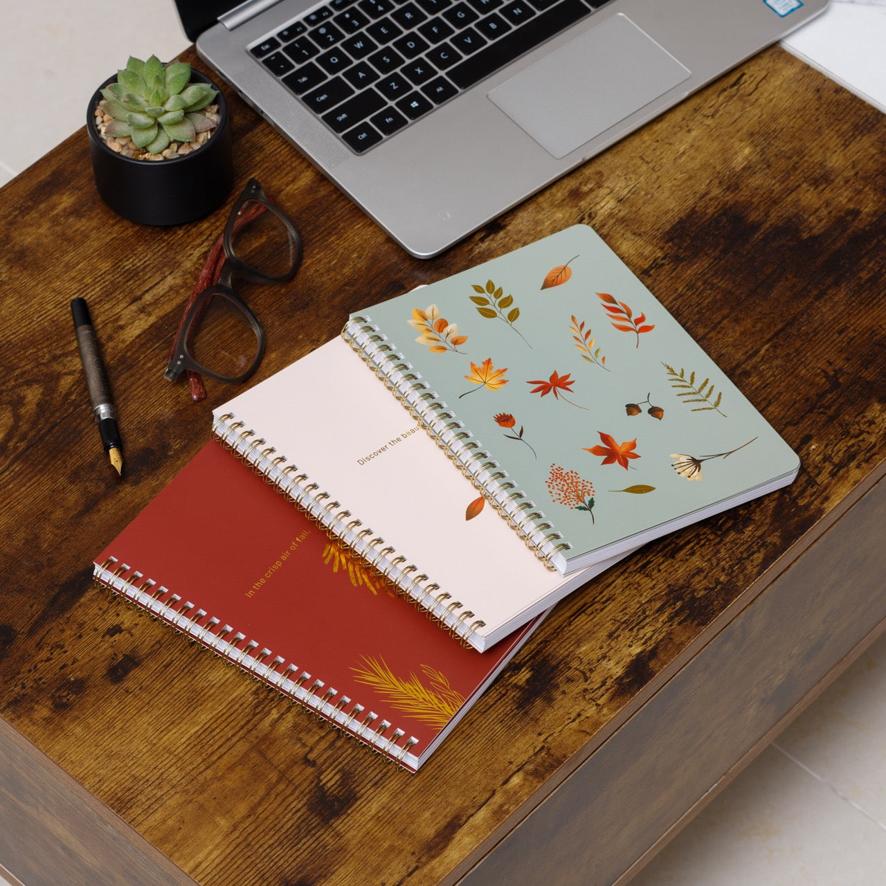 Three notebooks with floral designs on a wooden desk with a laptop and office supplies.