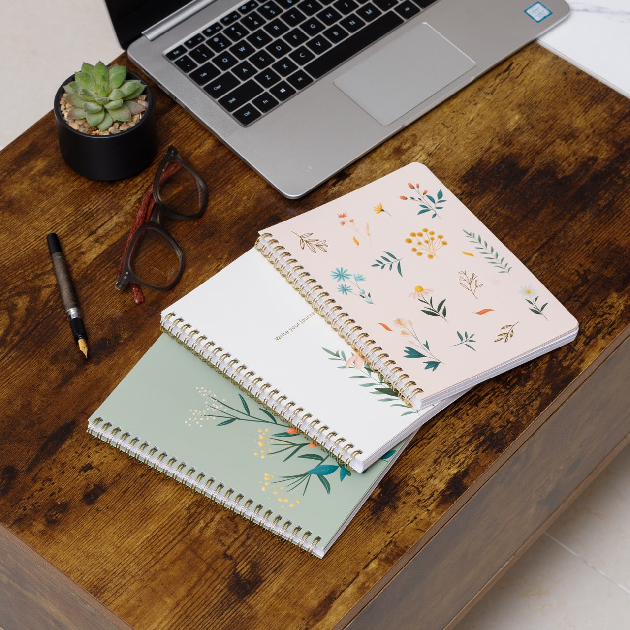 Two spiral-bound notebooks with floral designs on a wooden desk with a laptop and office supplies.