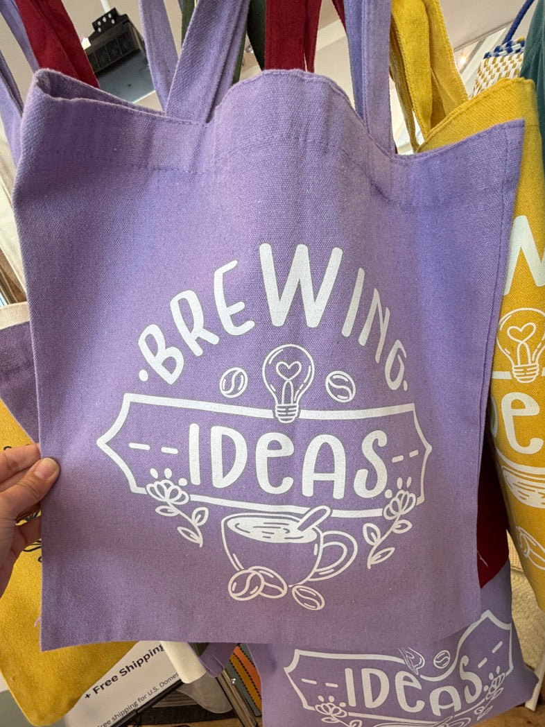 Purple tote bag with 'Brewing Ideas' text held by a hand in a store setting.