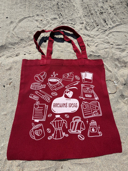 Red tote bag with white coffee-themed graphics on a sandy surface