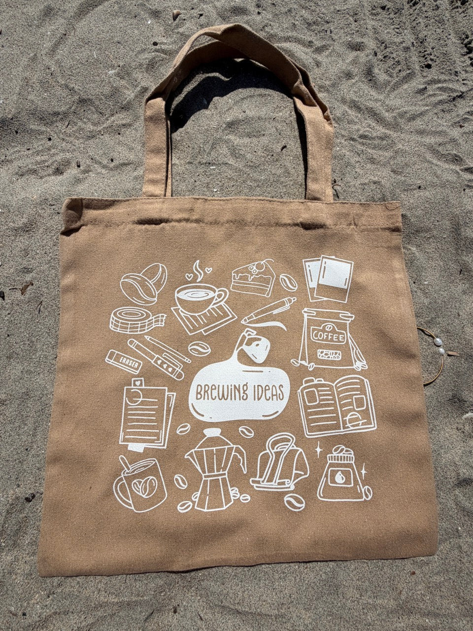 Brown tote bag with white coffee-themed illustrations on a sandy background