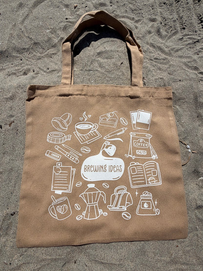 Brown tote bag with white coffee-themed illustrations on a sandy background