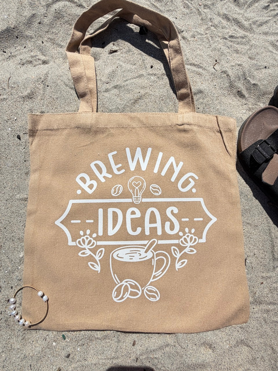 Beige tote bag with 'Brewing Ideas' text and graphic on sandy ground