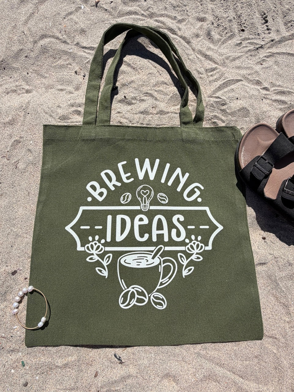 Green tote bag with 'Brewing Ideas' design on sandy ground