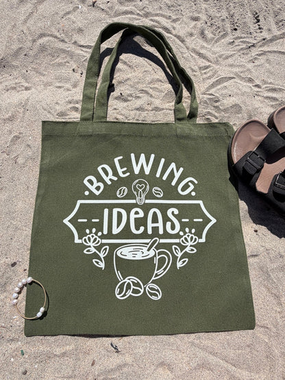 Green tote bag with 'Brewing Ideas' design on sandy ground