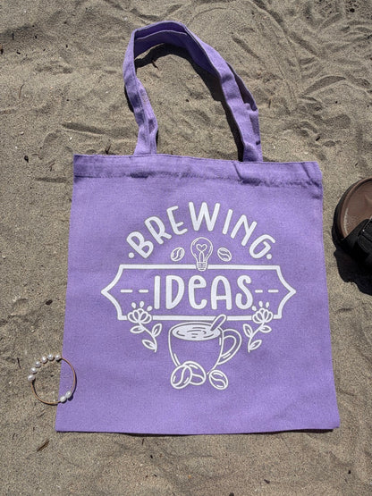 Purple tote bag with 'Brewing Ideas' text and graphics on a sandy surface