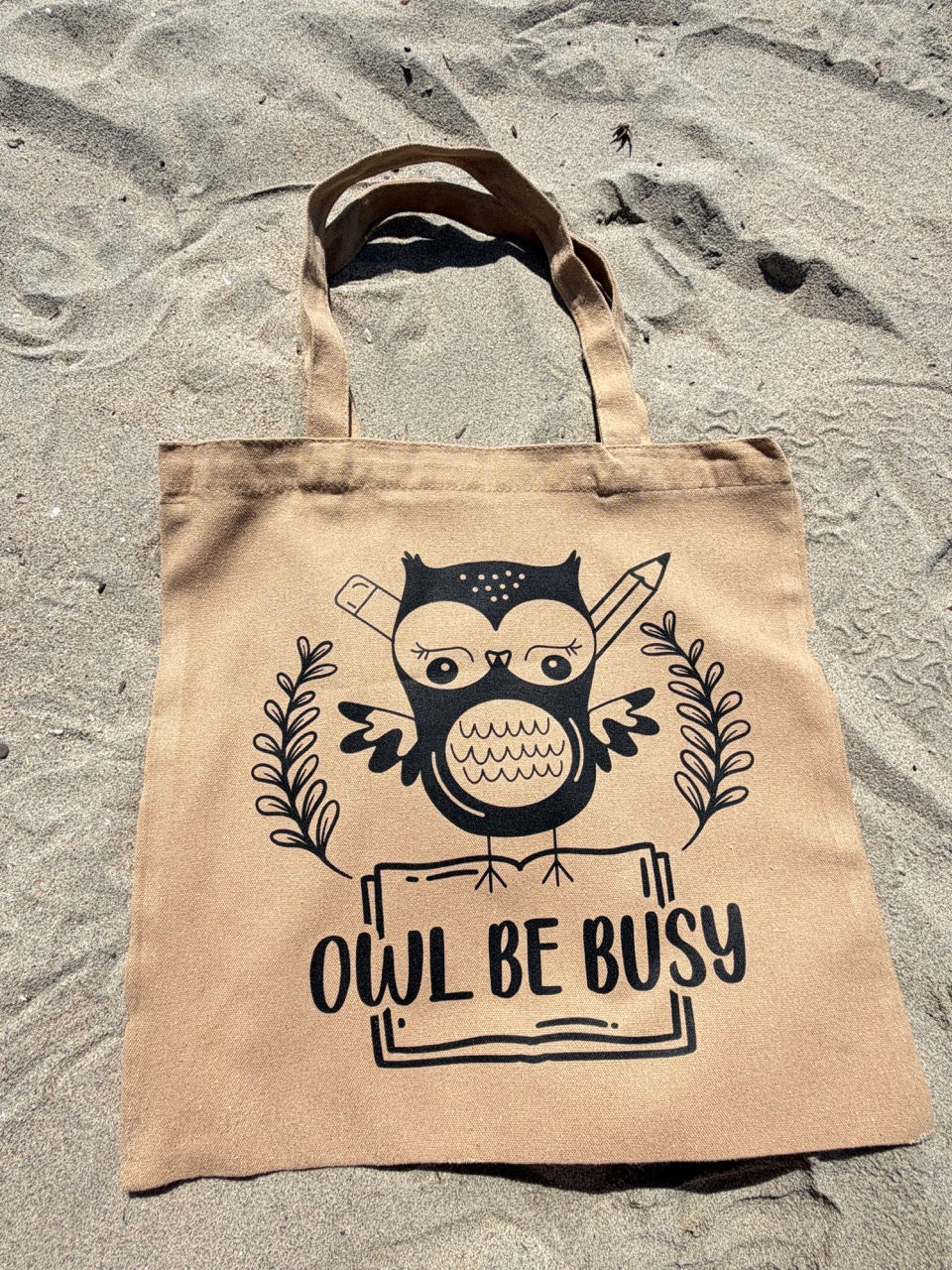 Beige tote bag with owl design and 'OWL BE BUSY' text on sandy surface