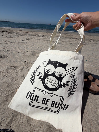 Tote bag with owl design and 'OWL BE BUSY' text held on a beach.