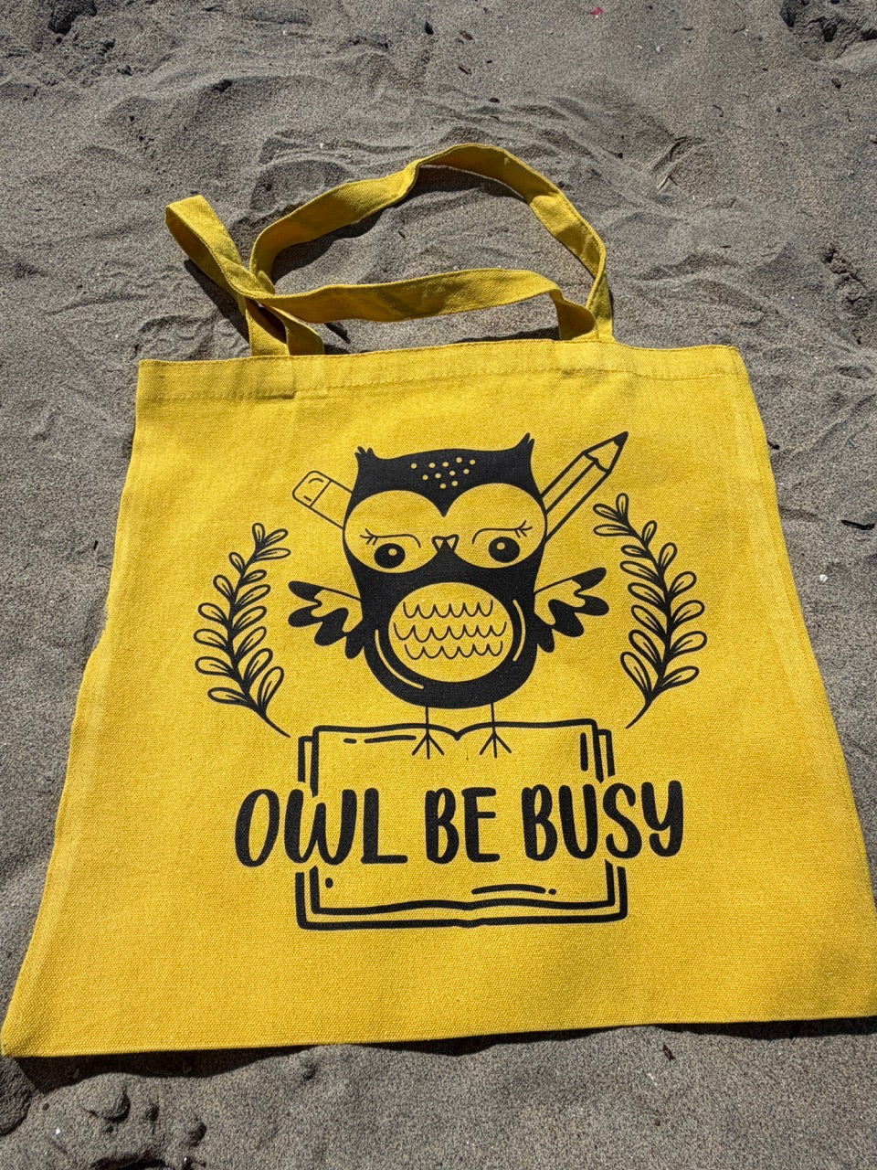 Yellow tote bag with owl design and 'OWL BE BUSY' text on sandy surface