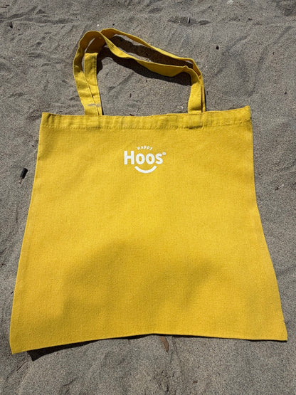 Yellow tote bag with "Happy Hoos' logo on a sandy surface