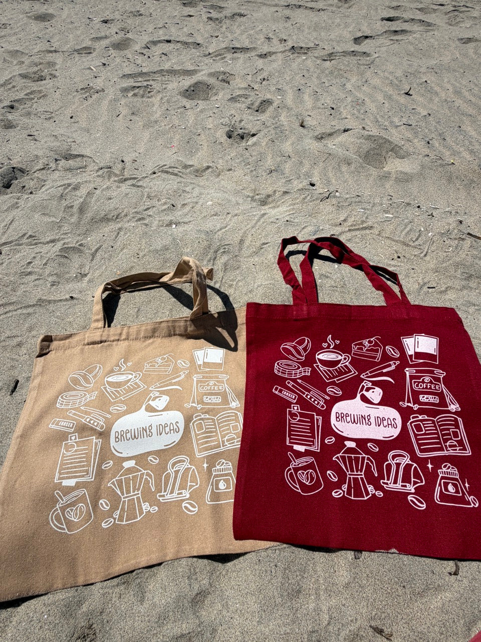 Two tote bags, one beige and one red, with coffee-themed designs on a stone surface.