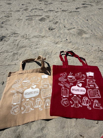 Two tote bags, one beige and one red, with coffee-themed designs on a stone surface.
