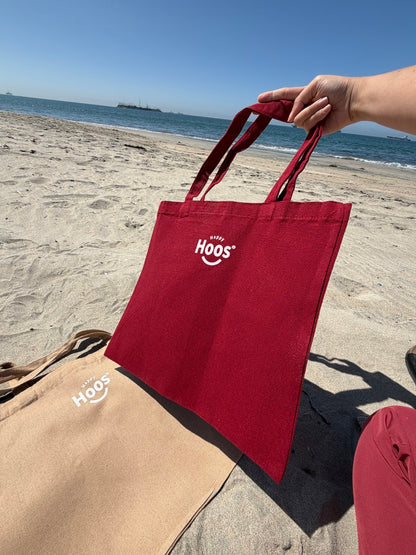 Red tote bag with 'HOOS' logo held by a person on a beach.