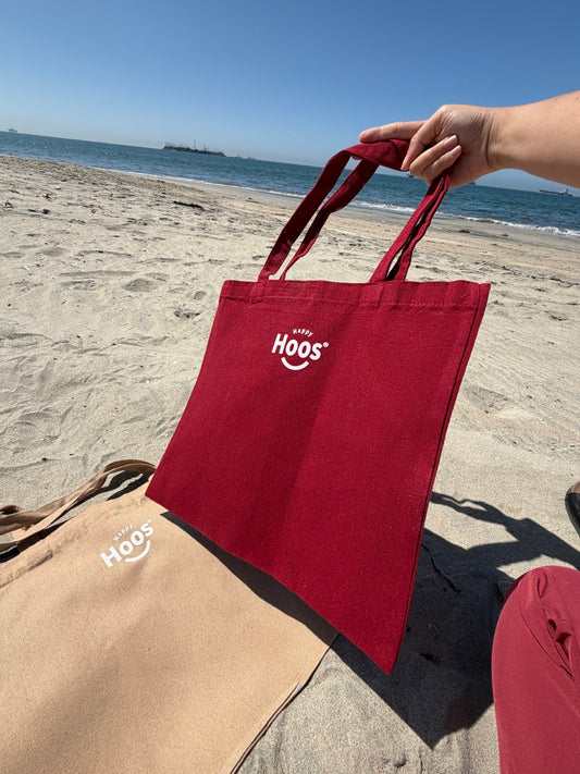 Red tote bag with 'HAPPY HOOS' logo held by a person on a beach.