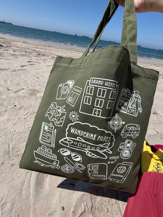 Green tote bag with white graphics held on a sandy beach about journaling and travellng 