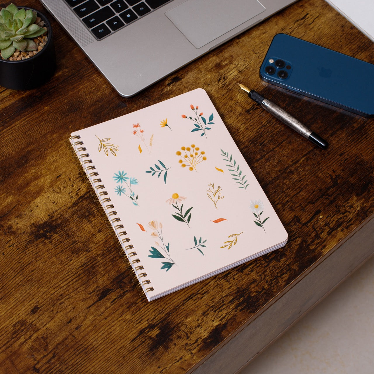 Notebook with floral pattern on a wooden desk with laptop, phone, and pen.