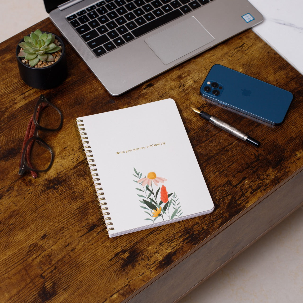 Notebook with floral design on a wooden desk with laptop, phone, and glasses.