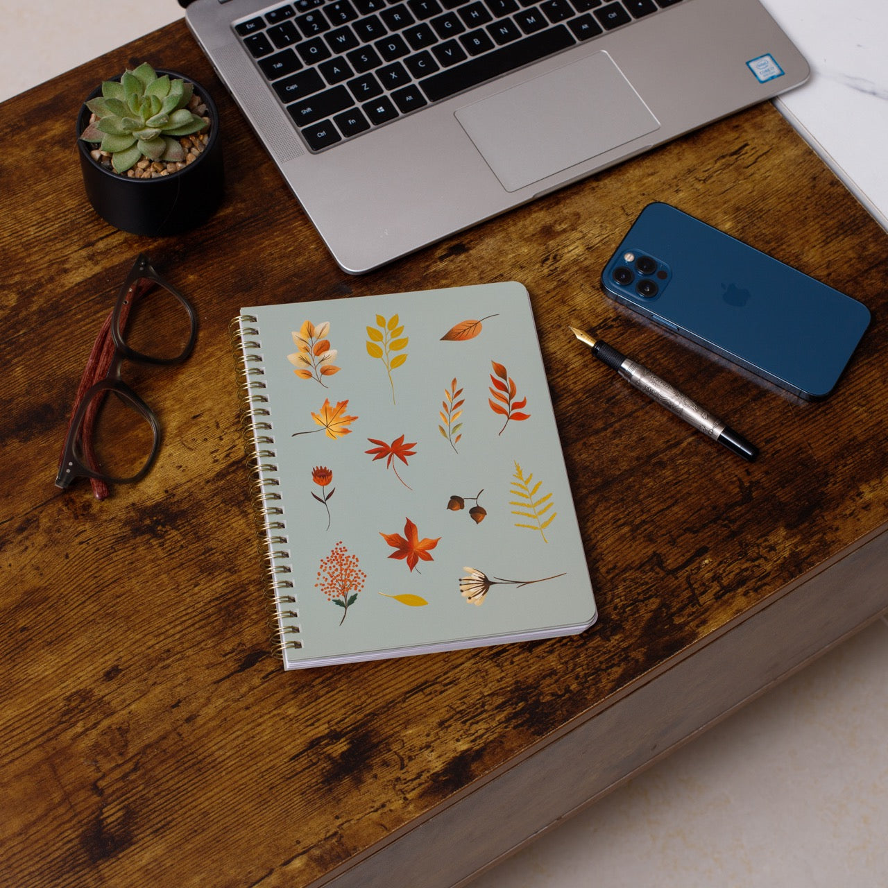 Notebook with autumn leaf design on a wooden desk with laptop, phone, and glasses.