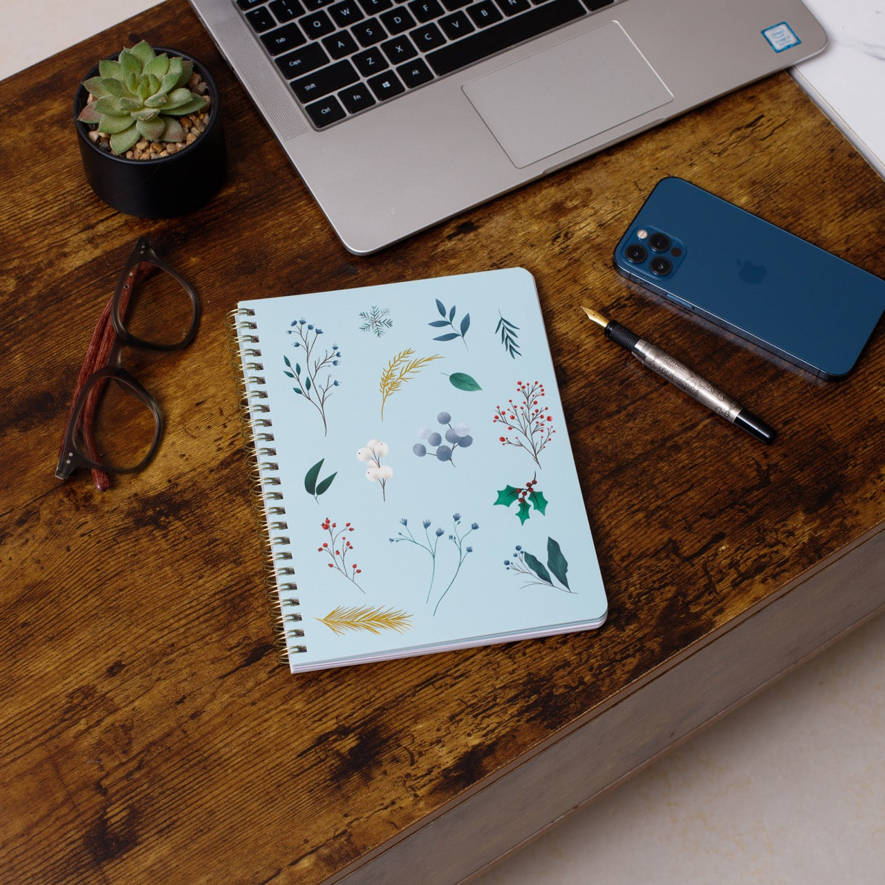 Notebook with floral design on a wooden desk with laptop, phone, and glasses.