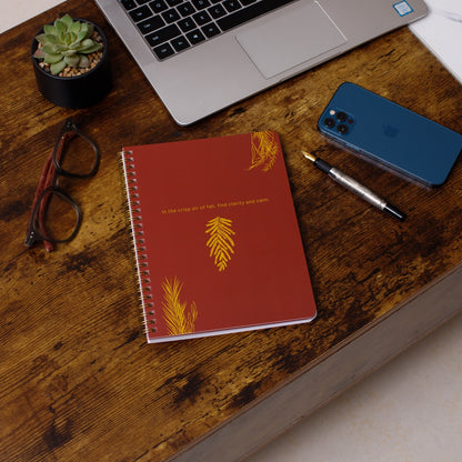Red notebook with yellow designs on a wooden desk with a laptop, phone, and glasses.