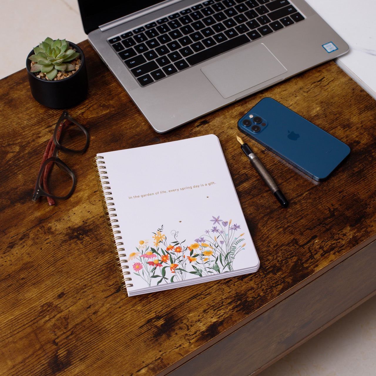 Laptop, notebook with floral design, smartphone, and glasses on a wooden desk.