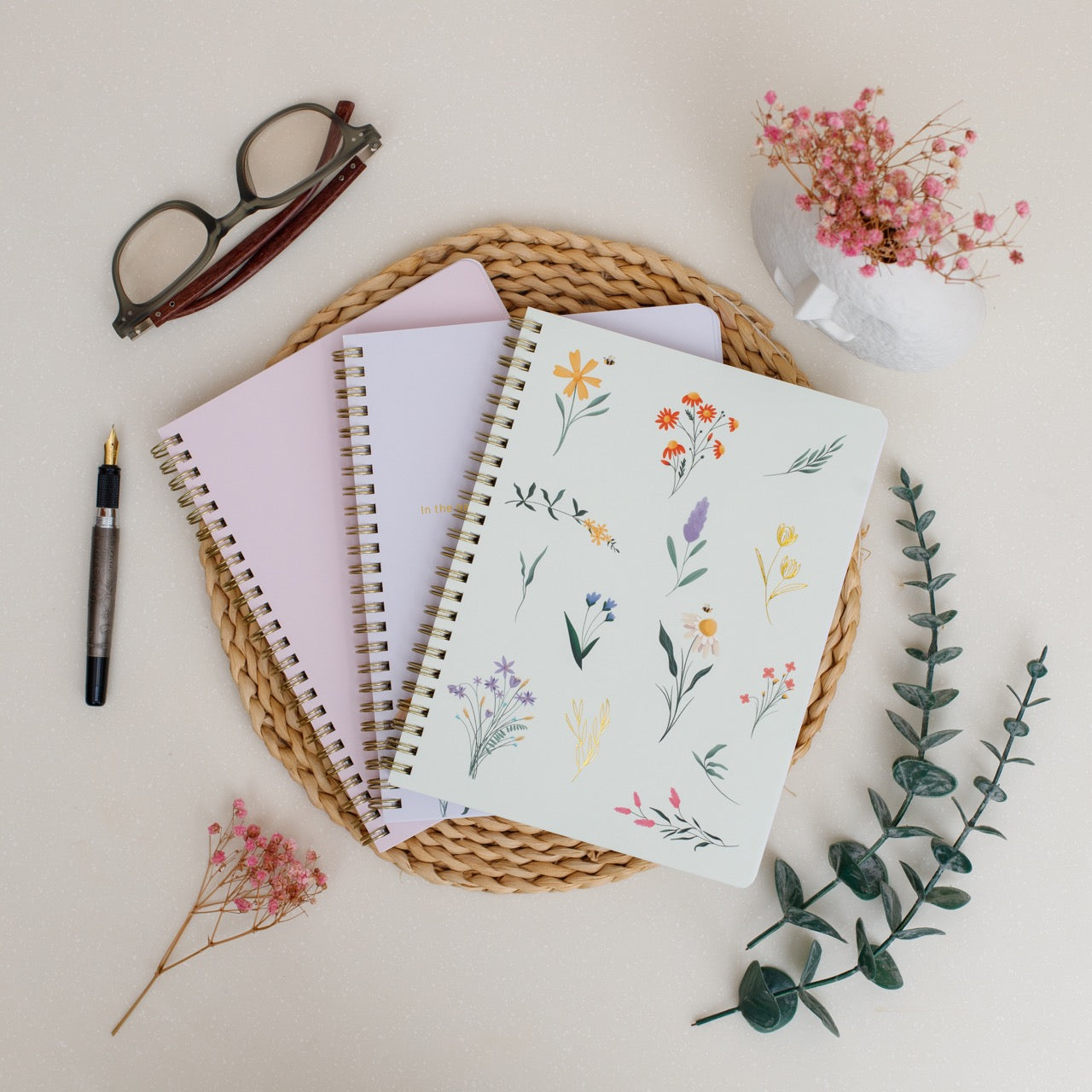 Set of notebooks with floral designs on a woven mat with a pen and glasses.