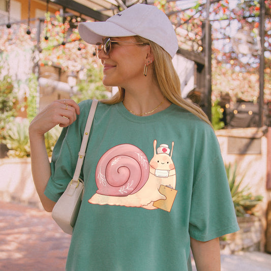 Woman wearing light green t-shirt with an illustration of snail holding a clipboard and wearing a nurse's hat and face mask.