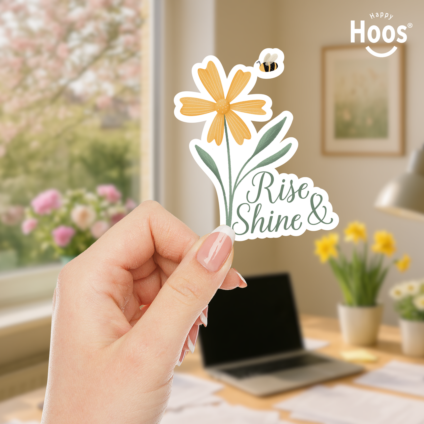 Hand holding a 'Rise and Shine' sticker with a yellow flower design against a blurred floral background.