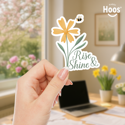 Hand holding a 'Rise and Shine' sticker with a yellow flower design against a blurred floral background.