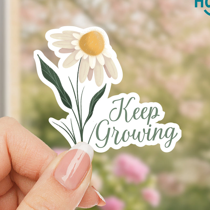 Hand holding a 'Keep Growing' sticker with a daisy design against a blurred floral background.