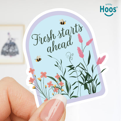 Hand holding a decorative sticker with floral design and 'Fresh starts ahead' text, with 'Happy Hoos' branding.