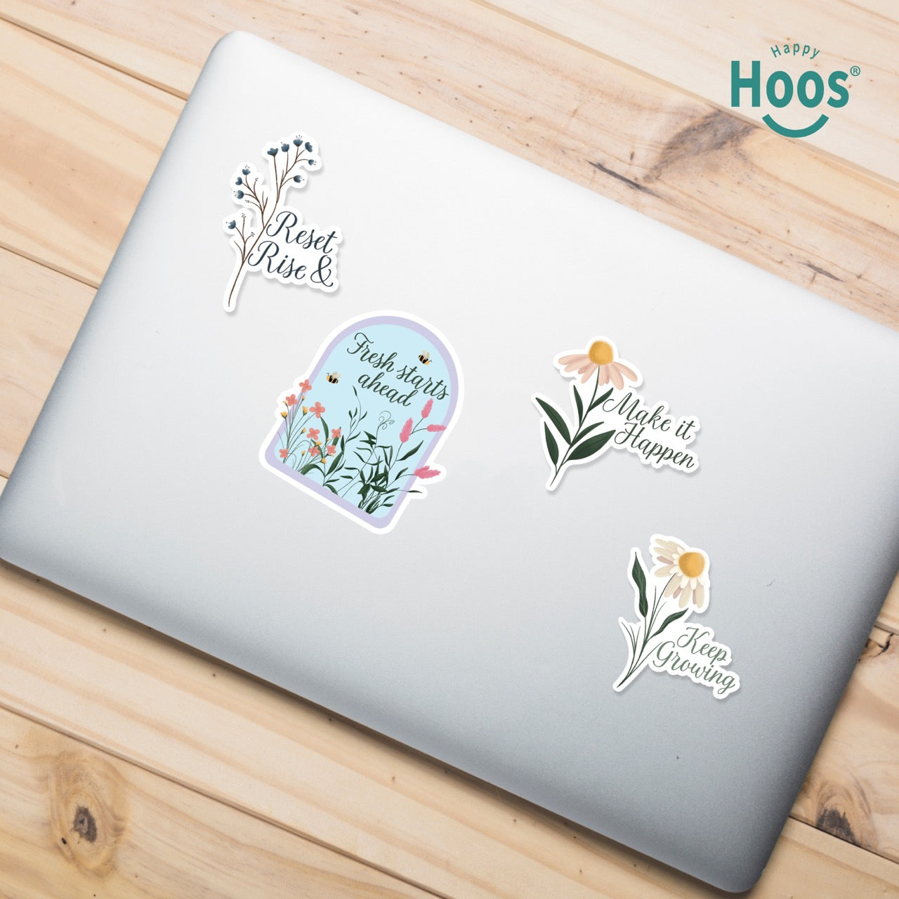 Laptop on a wooden surface with Motivation sticker pack with 4 floral design stickers  featuring the Hoos brand. The following text are presented in each sticker: Reset & Rise, Fresh Starts Ahead, make it Happen, Keep growing. 
