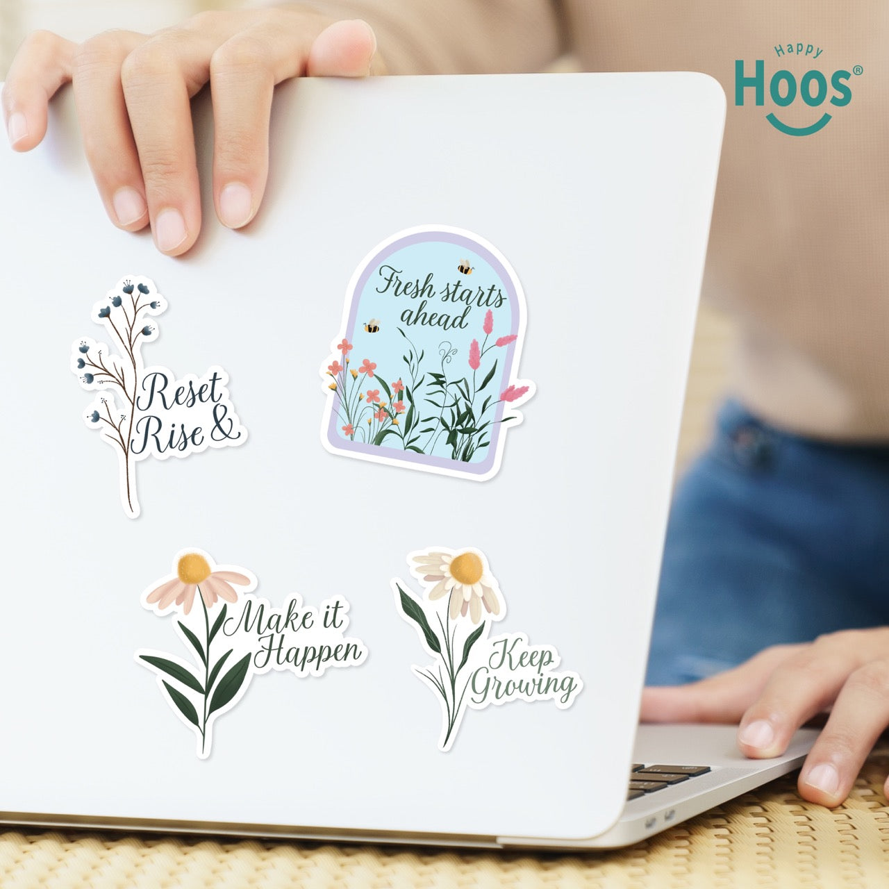 Laptop on a wooden surface with Motivation sticker pack with 4 floral design stickers  featuring the Hoos brand. The following text are presented in each sticker: Reset & Rise, Fresh Starts Ahead, make it Happen, Keep growing. 