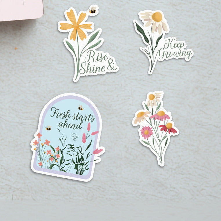 Four spring stickers on surface: daisy with bee and "Rise & Shine", daisy with "Keep Growing", garden dome with "Fresh starts ahead", colorful wildflower bouquet