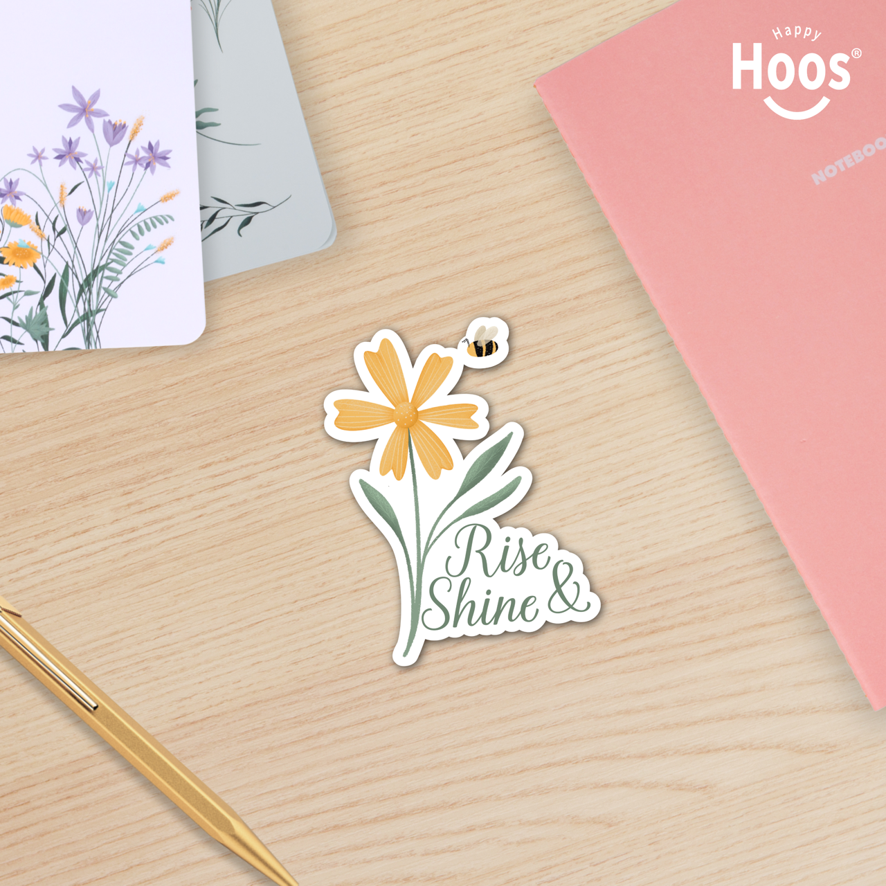 Sticker with a flower design and 'Rise & Shine' text on a wooden surface with stationery items.