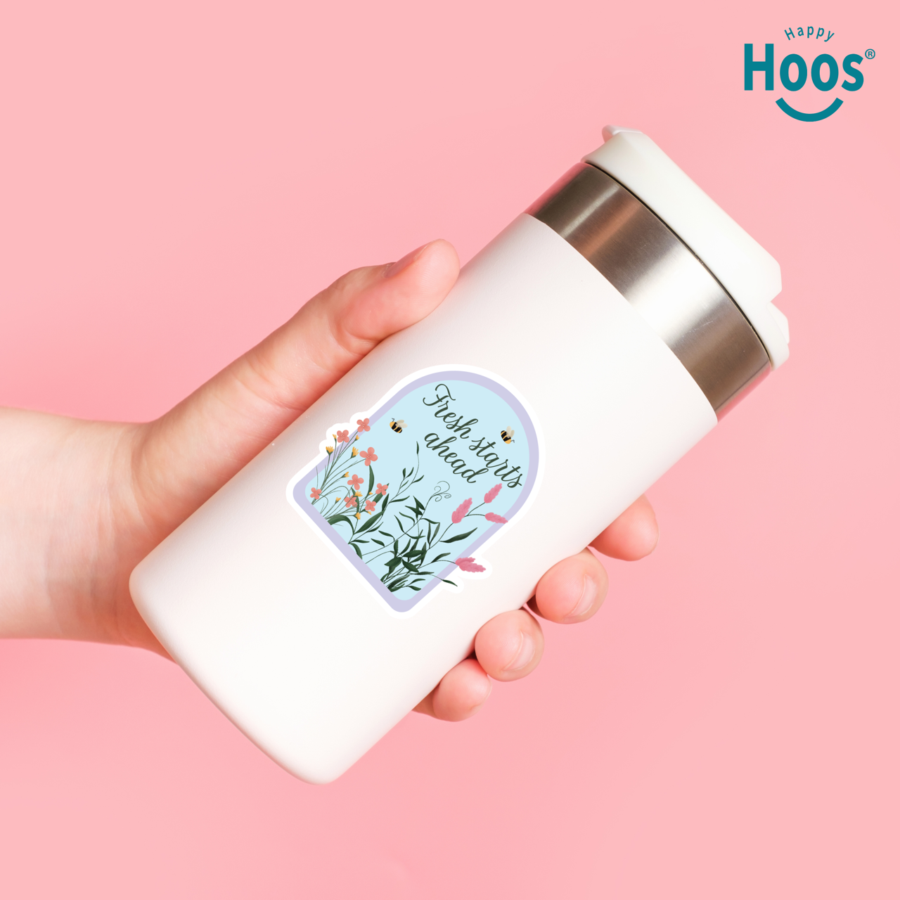 Hand holding a white thermos with a floral label on a pink background, featuring the 'Happy Hoos' brand.