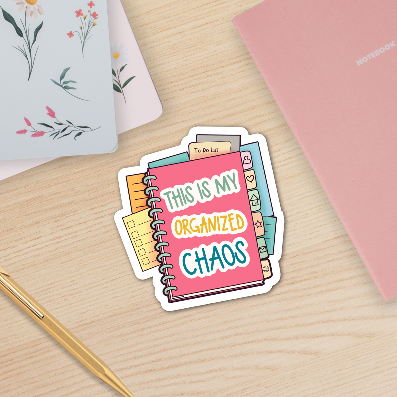 Sticker with 'This is my organized chaos' text on a desk with notebooks and a pen.