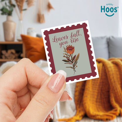hand holding 
Fall & Rise sticker designed as a vintage postage stamp with burgundy perforated border, sage green background, featuring autumn botanicals with orange flowers and brown foliage, and burgundy script lettering reading "Leaves fall, you rise" at the top, measuring 2.5 by 3.3 inches