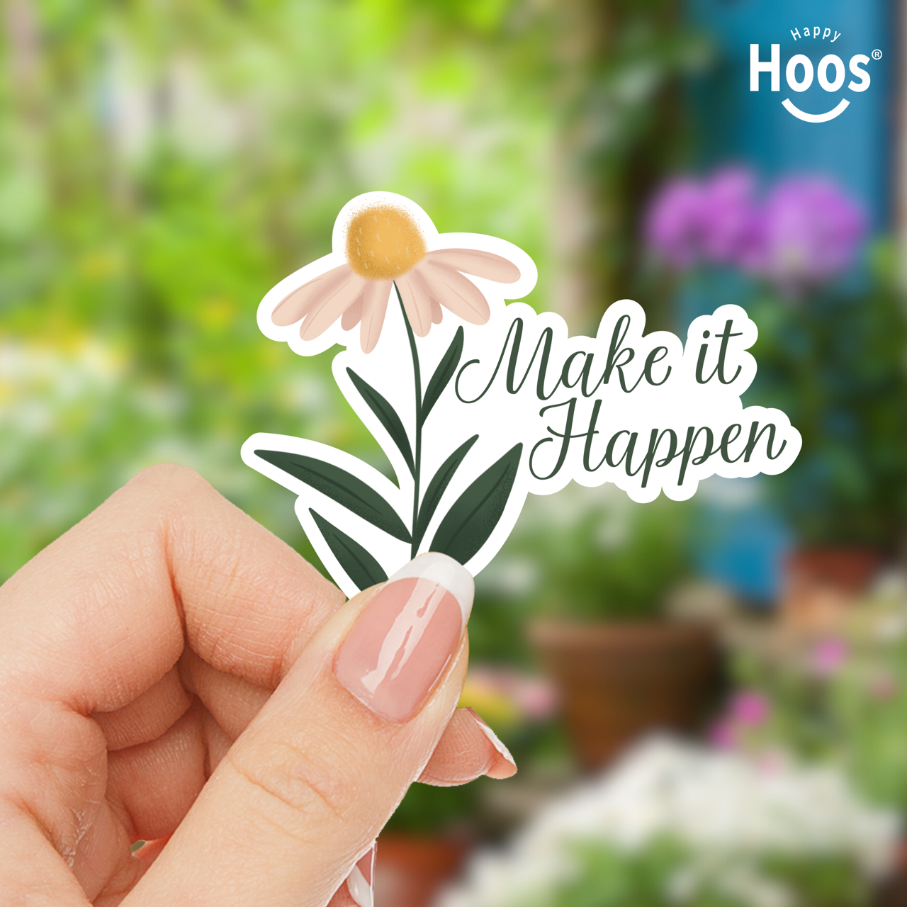 Hand holding a sticker with a flower and 'Make it Happen' text, with a blurred garden background.