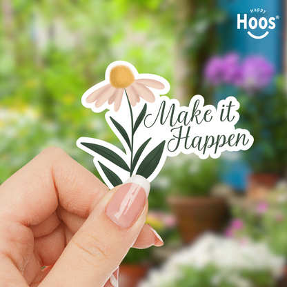 Hand holding a sticker with a flower and 'Make it Happen' text, with a blurred garden background.