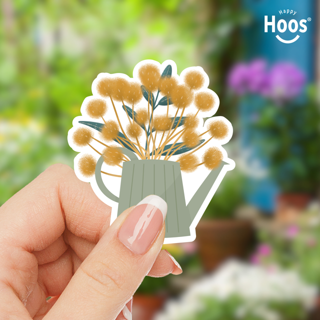 Hand holding a sticker of a watering can with flowers against a garden background