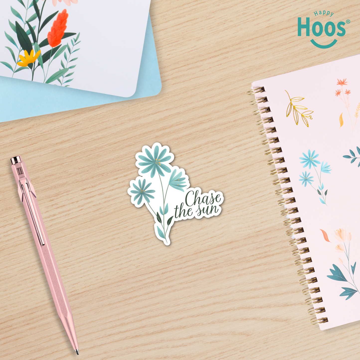 Sticker with floral design and 'Chase the sun' text on a wooden surface with a pen and notebook.