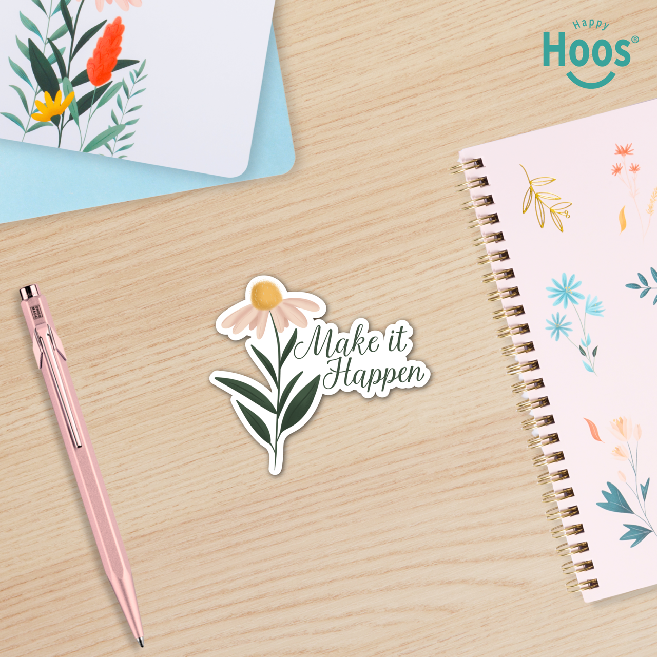 Sticker with floral design and 'Make it Happen' text on a wooden surface with a pen and notebook.