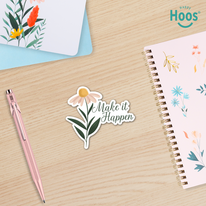 Sticker with floral design and 'Make it Happen' text on a wooden surface with a pen and notebook.
