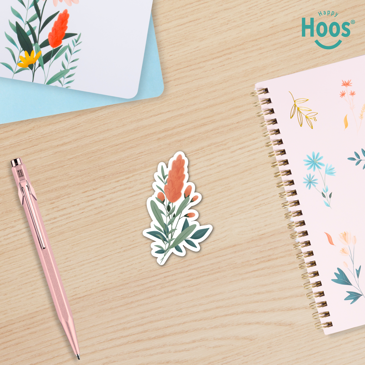 Floral sticker on a wooden surface with a pen and notebook, featuring the brand 'Happy Hoos'.