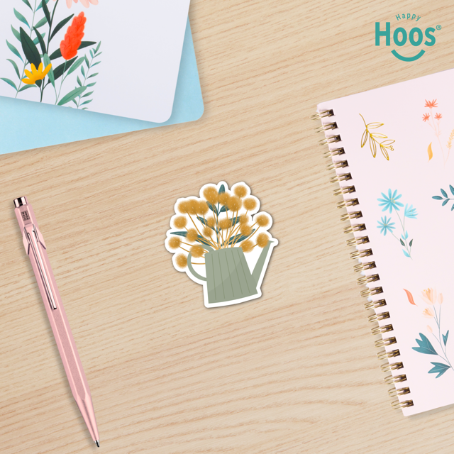 Sticker of a watering can with flowers on a wooden surface next to a pen and notebook.