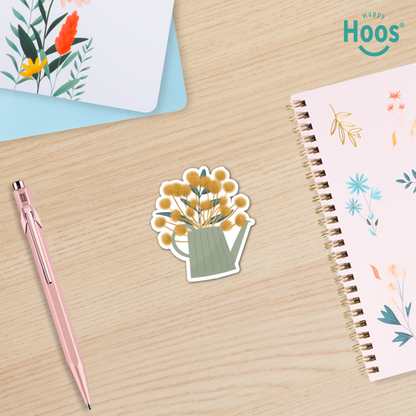 Sticker of a watering can with flowers on a wooden surface next to a pen and notebook.
